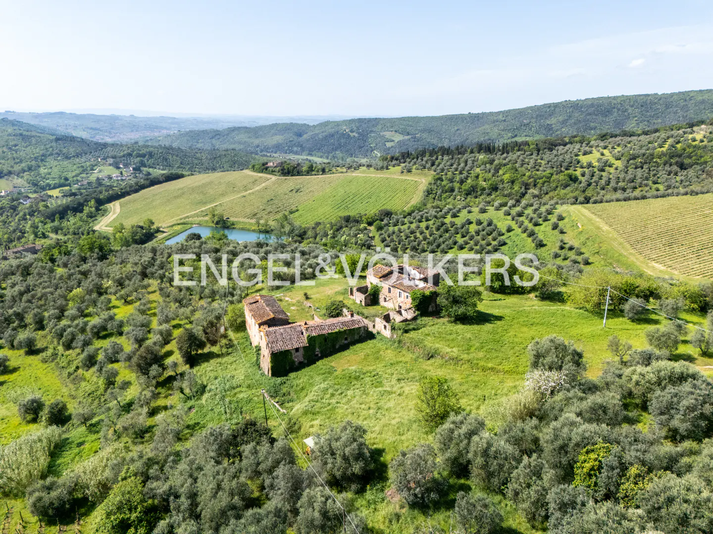 Aerial view of a Tuscan villa with a red tile roof, surrounded by green hills, olive groves, and a small lake.