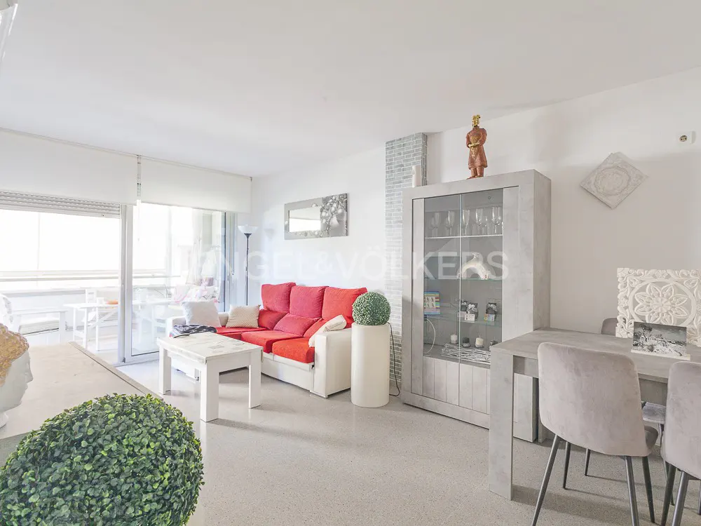 Bright living room with a red sofa, white coffee table, and gray dining set. A glass cabinet displays decorative items. Balcony access visible.