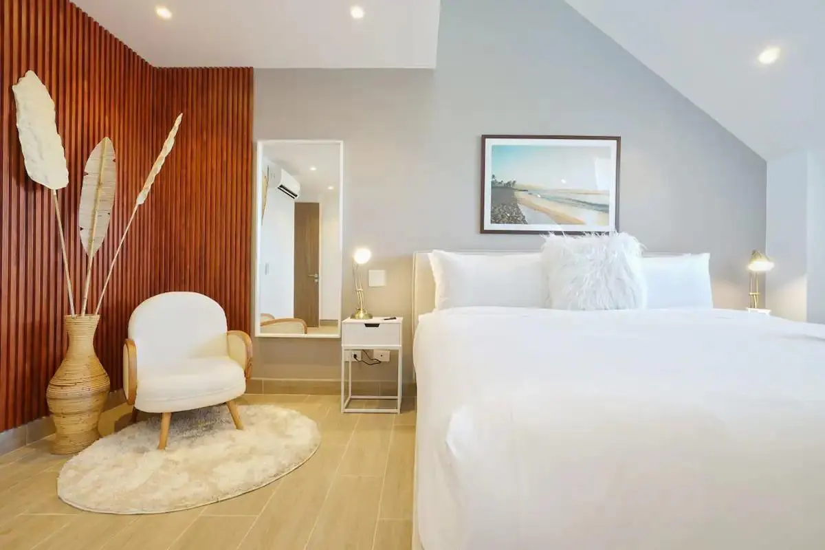 Bright bedroom with white bed, chair, and rug. Wood-paneled wall with vase and dried leaves. Beach art and mirror add to the room's decor.