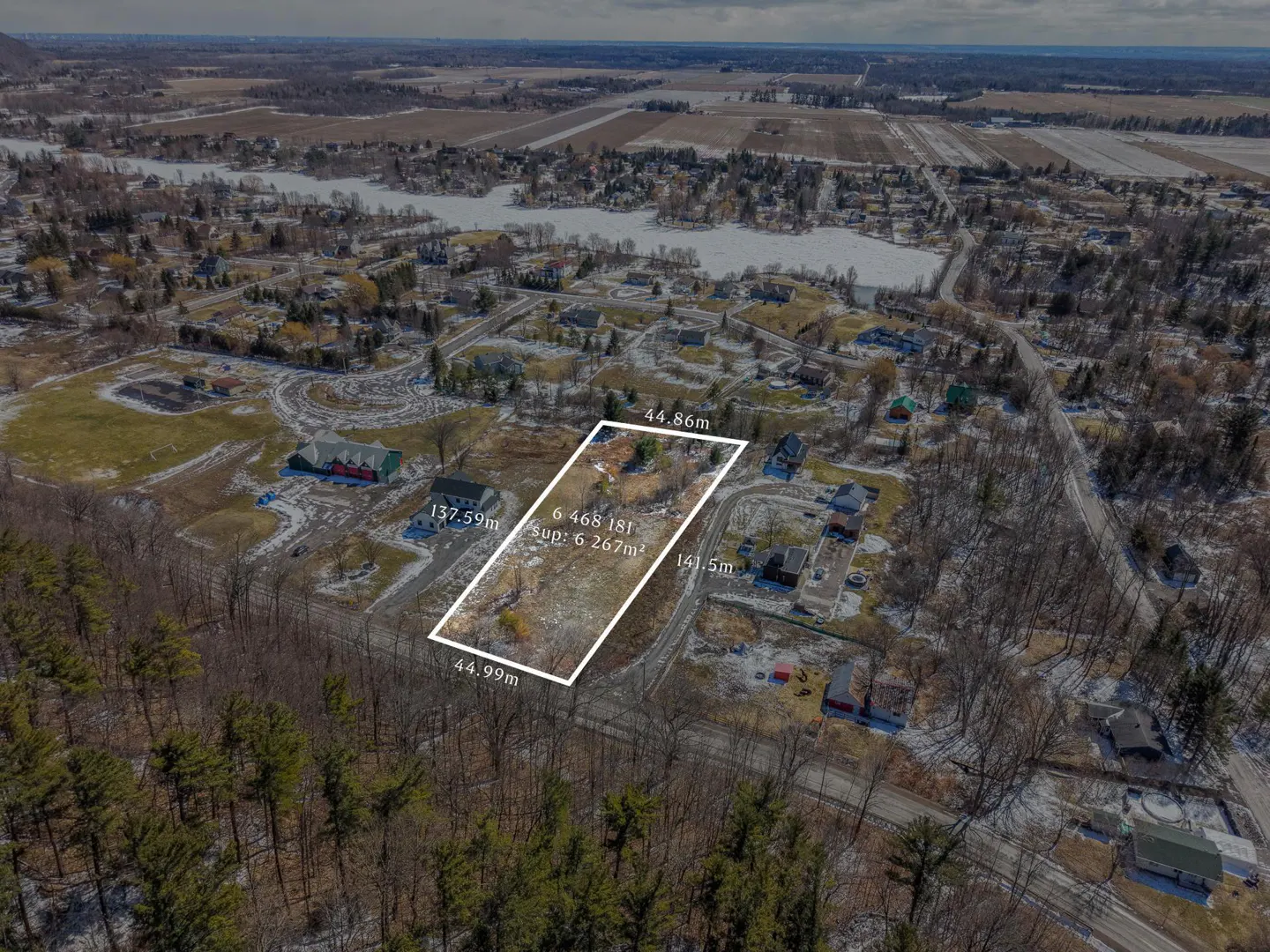 Aerial view of a vacant lot outlined in white, surrounded by trees and houses in a rural area with a lake in the background.