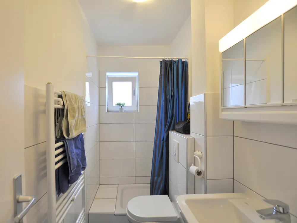 Bright bathroom with white tiles, a blue striped shower curtain, and a mirrored cabinet above a white sink. A towel rack holds folded towels.