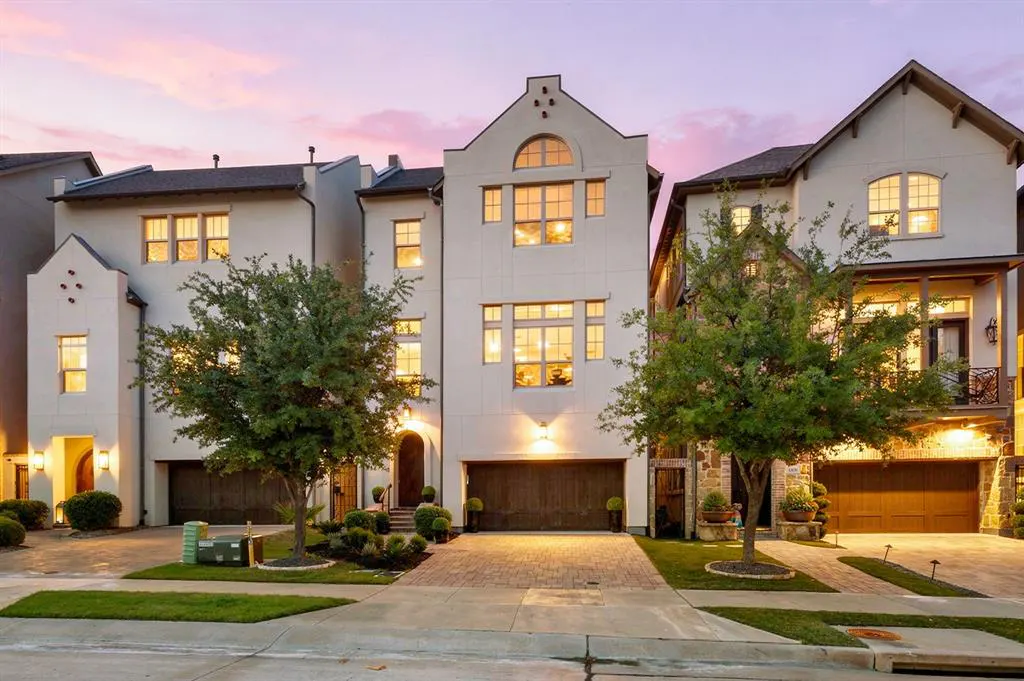 Three-story white townhouses with dark wood garage doors and green trees in front. Pink and purple sky.