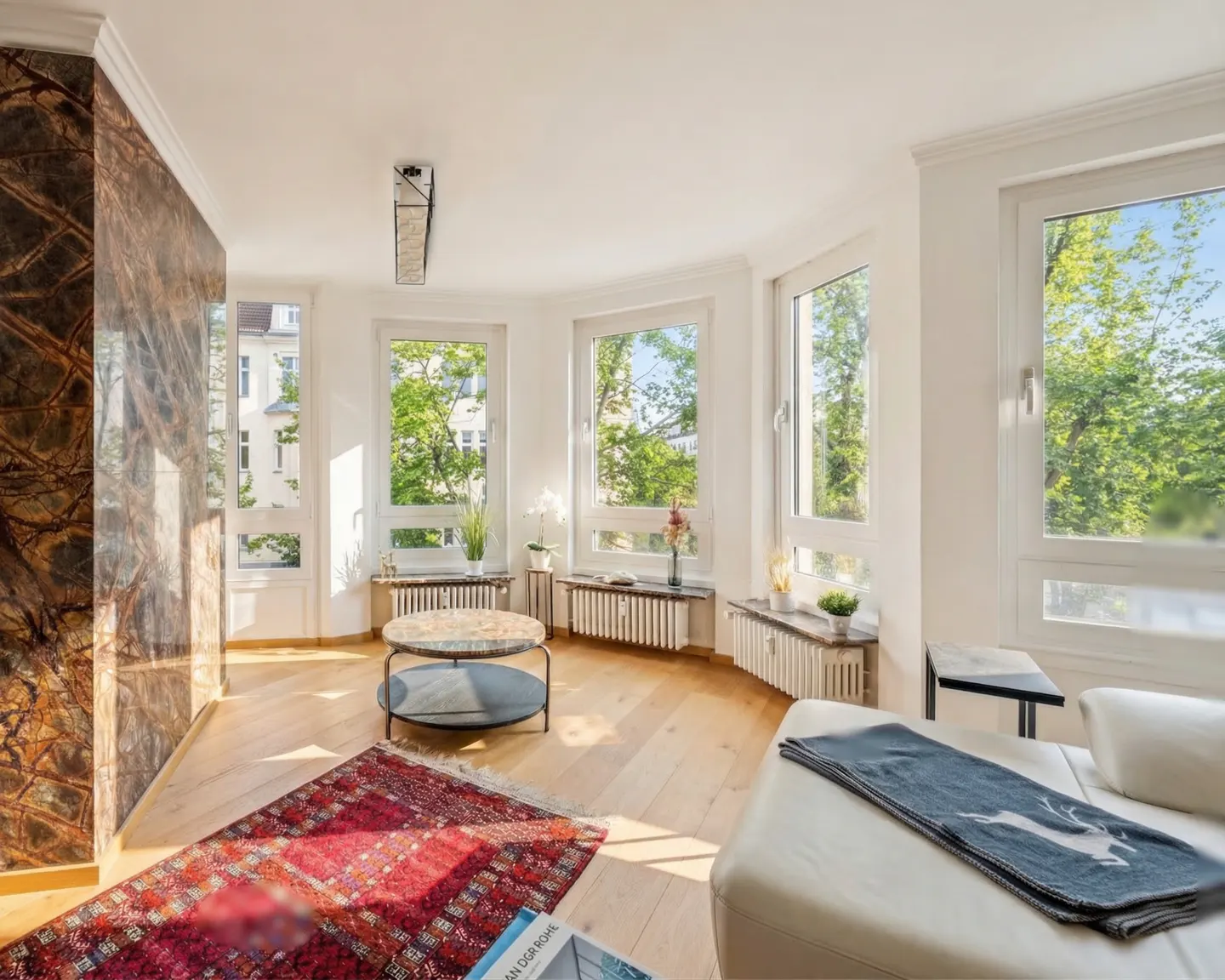Bright living room with wood floors, white walls, and large windows overlooking green trees. A red rug sits near a marble-accented wall and a white couch.