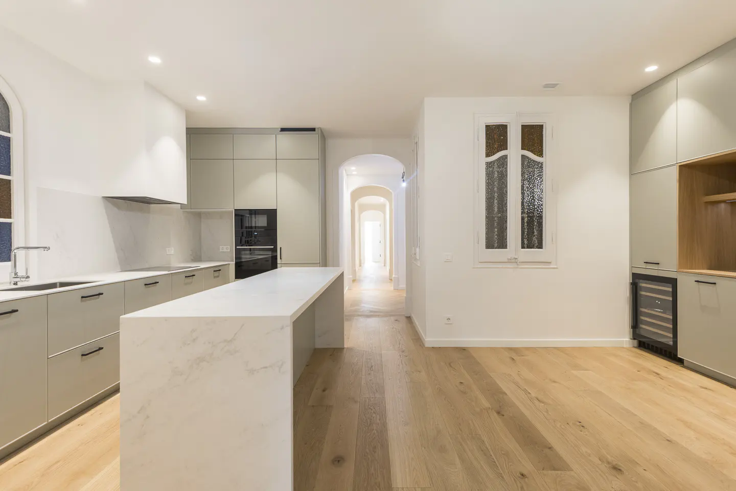 Bright, modern kitchen with light wood floors, gray cabinets, and a white marble island. A hallway with arched doorways extends into the distance.