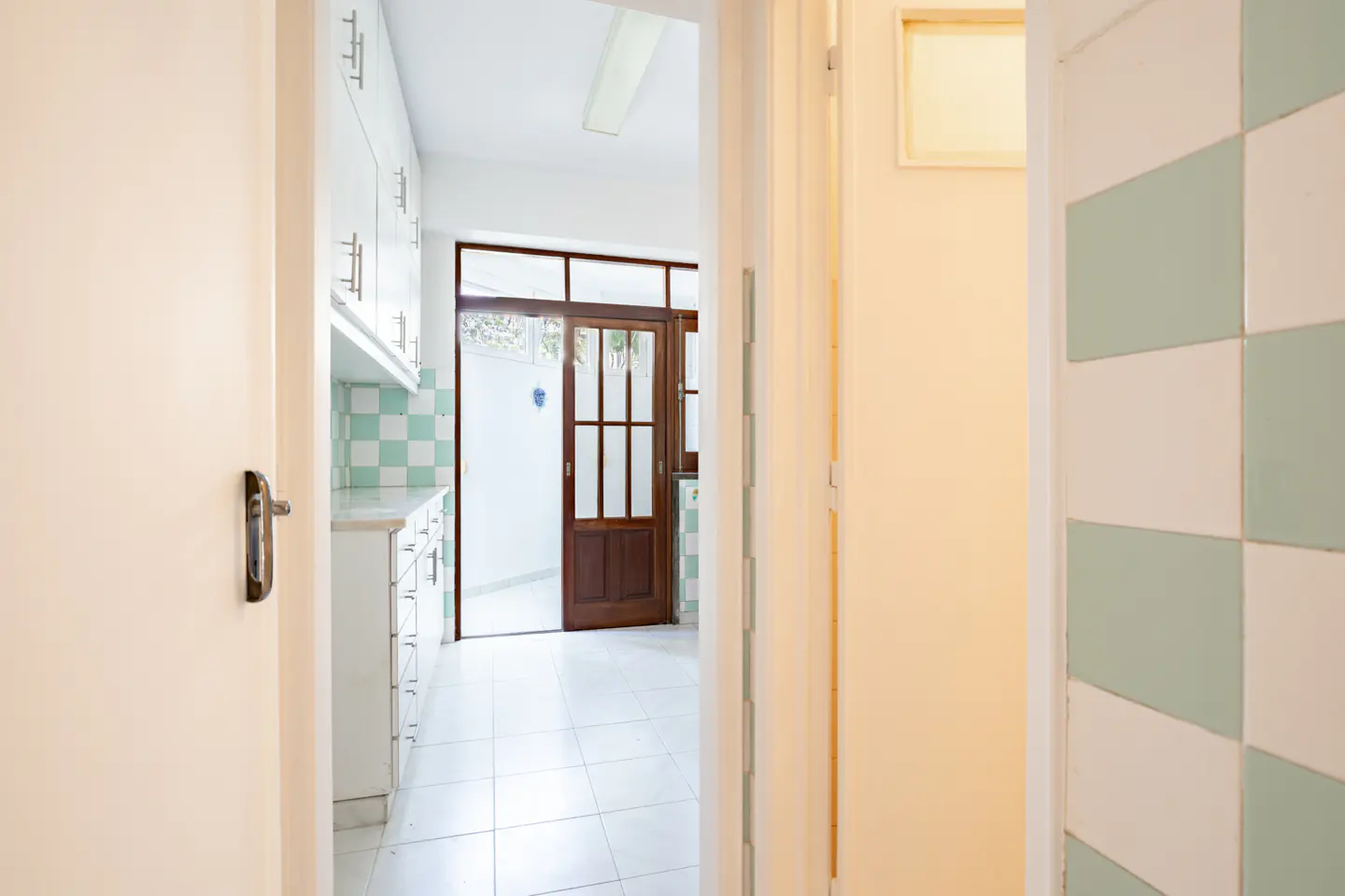 View through a doorway into a kitchen with white cabinets, green and white checkered tiles, and a wooden glass-pane door.