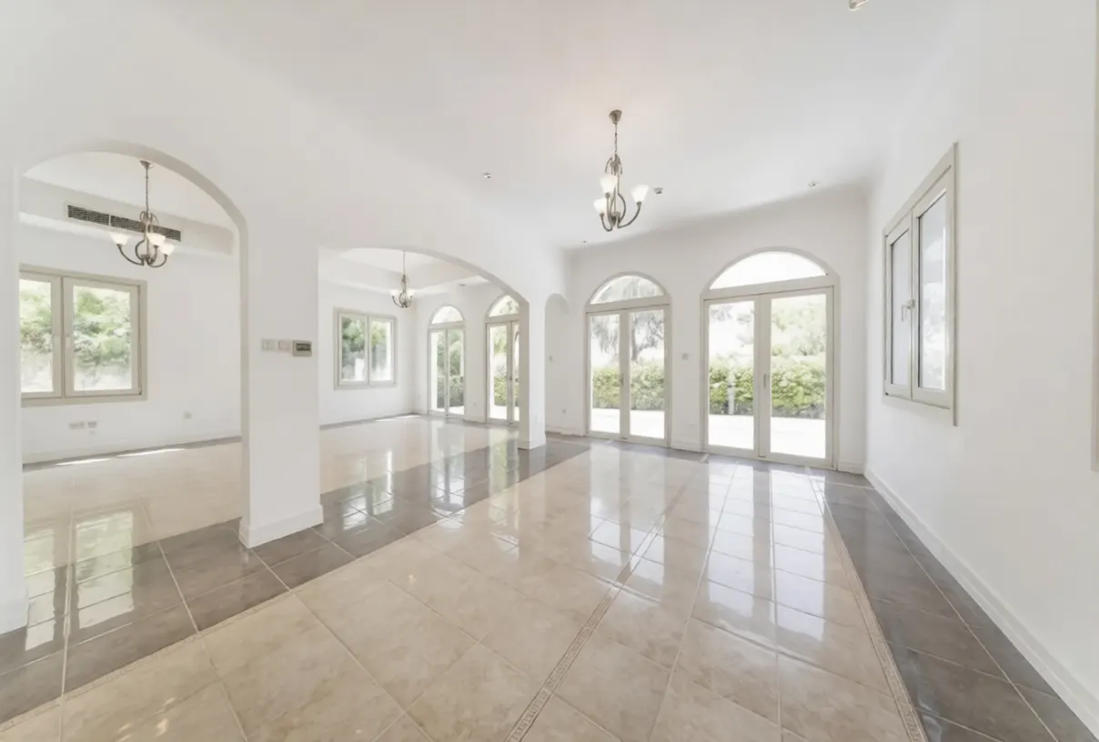 Bright, empty room with white walls, arched doorways, and tile floors. Chandeliers hang from the ceiling. Large windows offer a view of greenery.