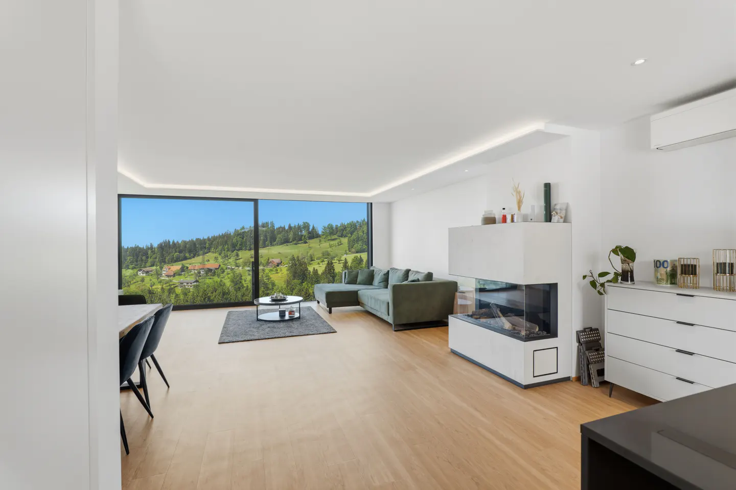 Bright living room with wood floors, a green sectional sofa, and a modern white fireplace. Large window with a view of green hills.