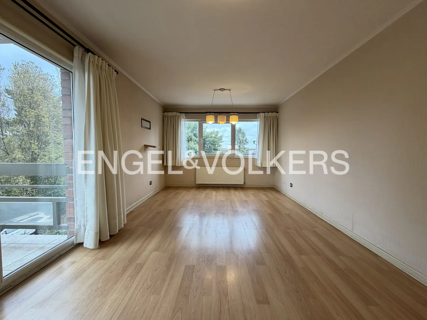 Empty living room with wood floors, beige walls, and white curtains. A balcony is on the left, and a window is at the back.
