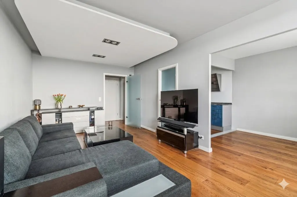 Living room with gray sectional sofa, wood floors, and a black TV on a stand. A kitchen is visible through an opening in the wall.