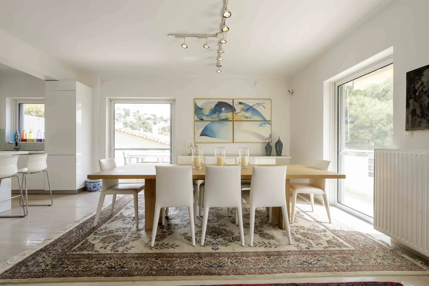 Bright dining room with a light wood table, white chairs, and a patterned rug. Artwork and natural light fill the space.
