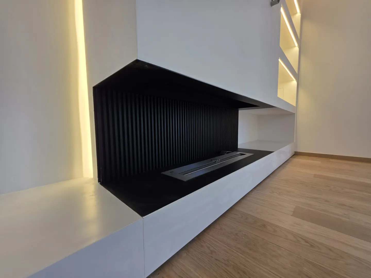 Modern white fireplace with black slatted back, integrated lighting, and light wood flooring in a minimalist room.