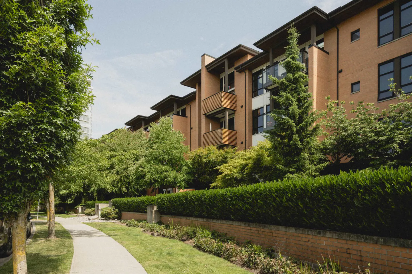 A modern tan brick apartment building with wooden balconies, surrounded by lush green trees and a sidewalk.