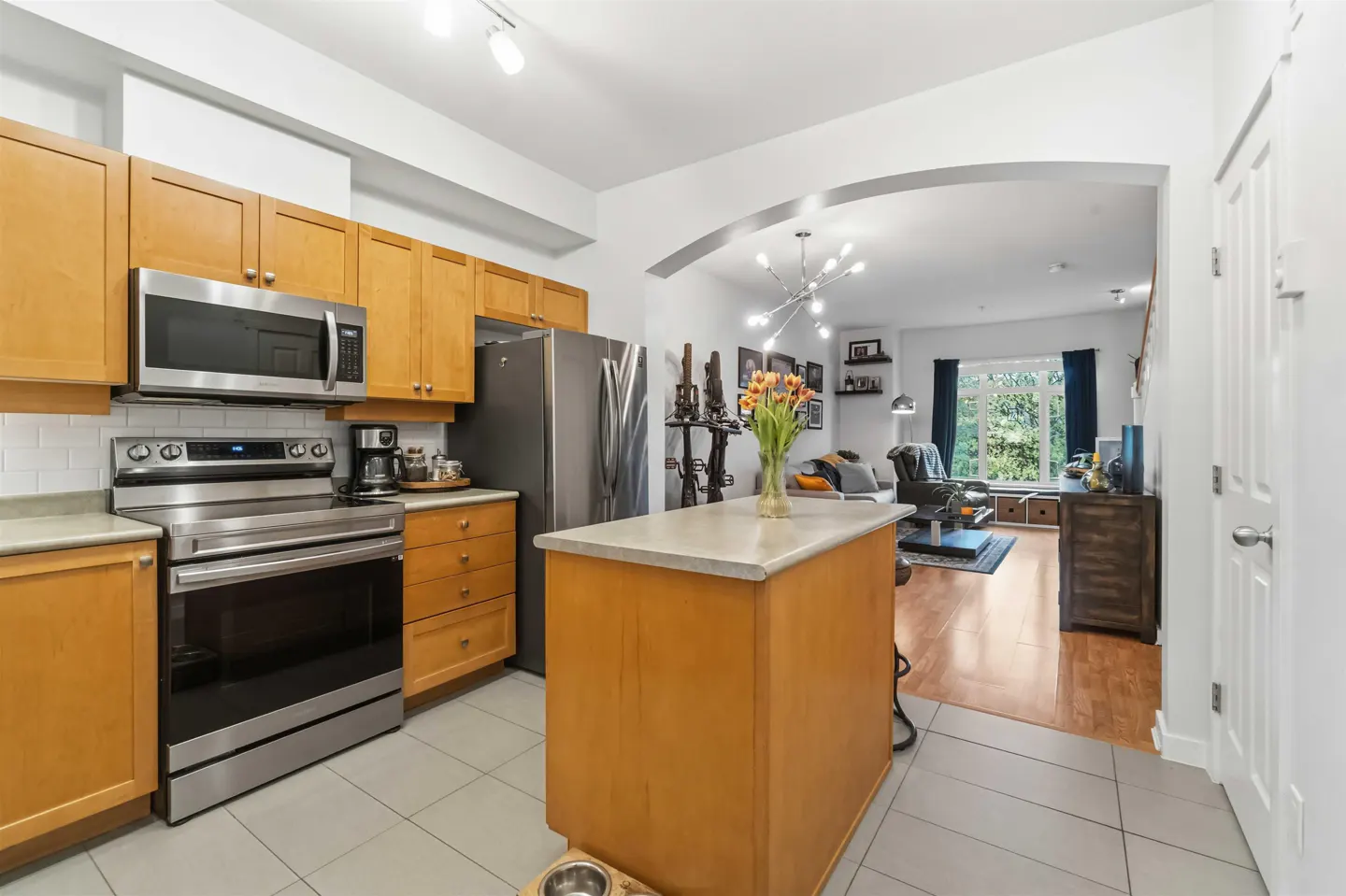 A bright, modern kitchen with light wood cabinets, stainless steel appliances, and an island, flowing into a living room with an arched doorway.