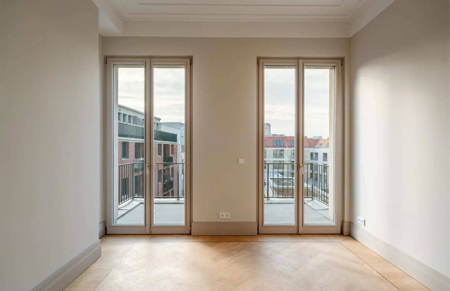 Empty room with light beige walls, herringbone wood floors, and two glass balcony doors overlooking city buildings.