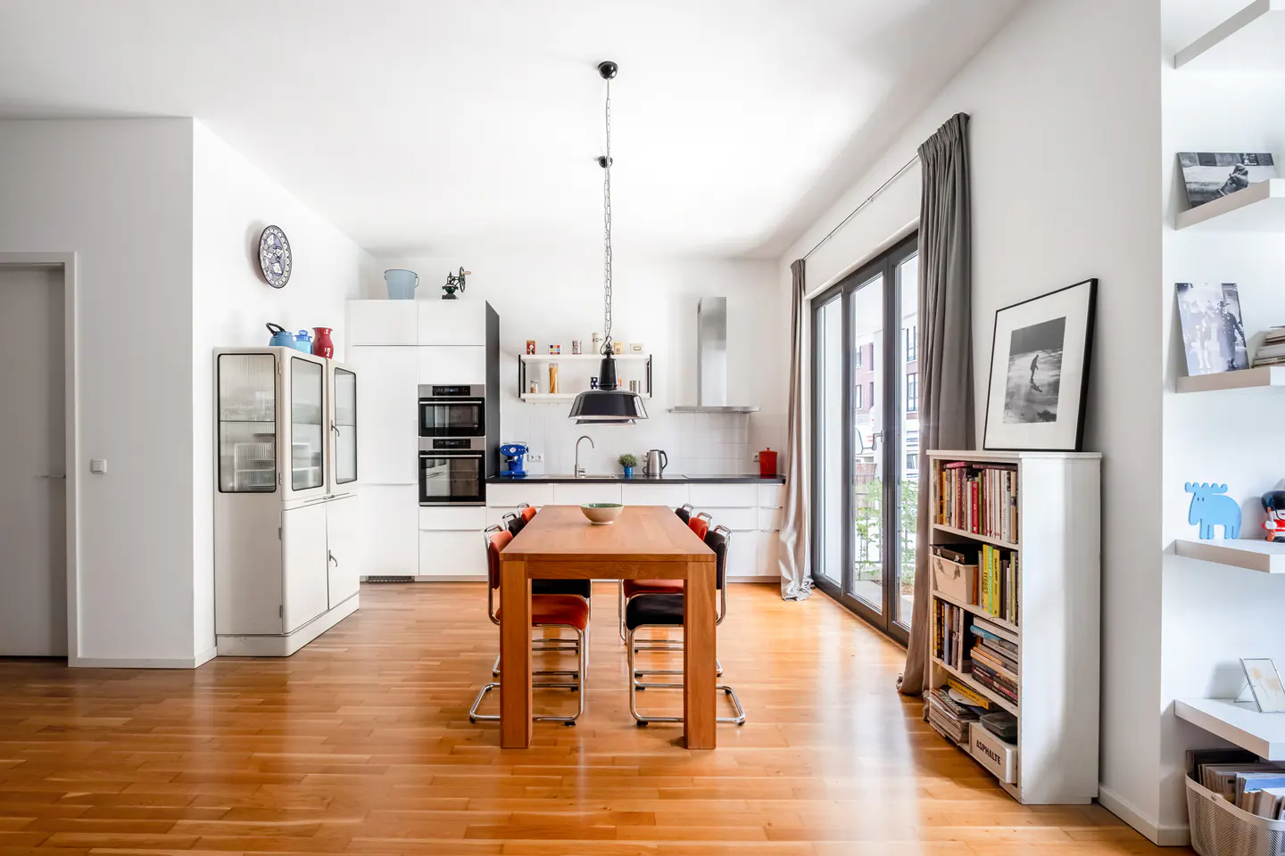 Modern kitchen with white cabinets, wooden dining table, chairs, and a bookshelf, featuring hardwood floors and glass doors.
