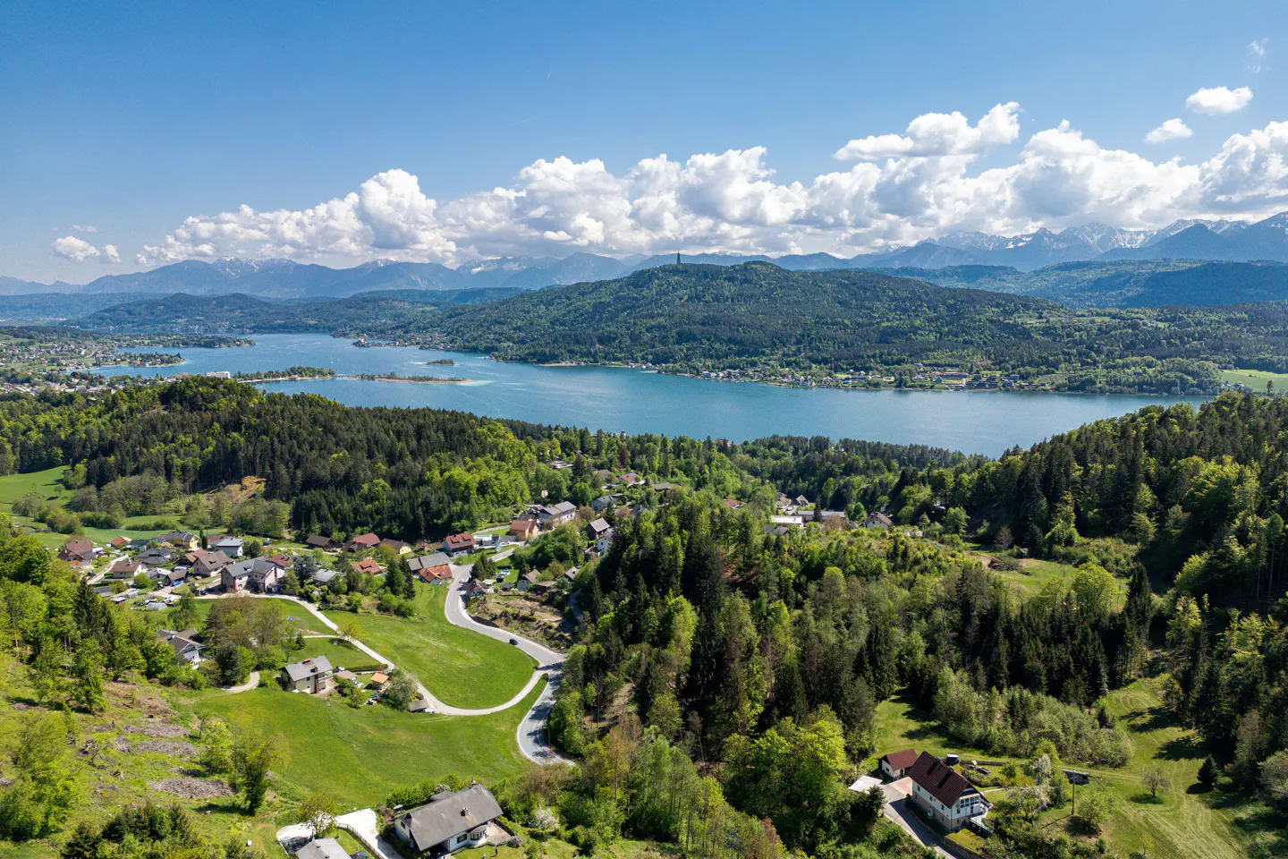 Aerial view of a blue lake surrounded by green hills, forests, and houses under a blue sky with white clouds. Snow-capped mountains are visible in the distance.
