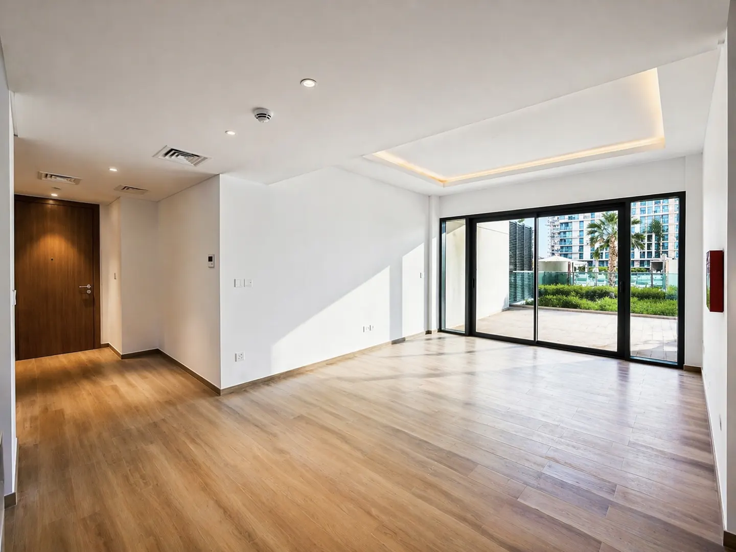 Bright, empty room with wood floors, white walls, and a large sliding glass door to a patio with greenery.