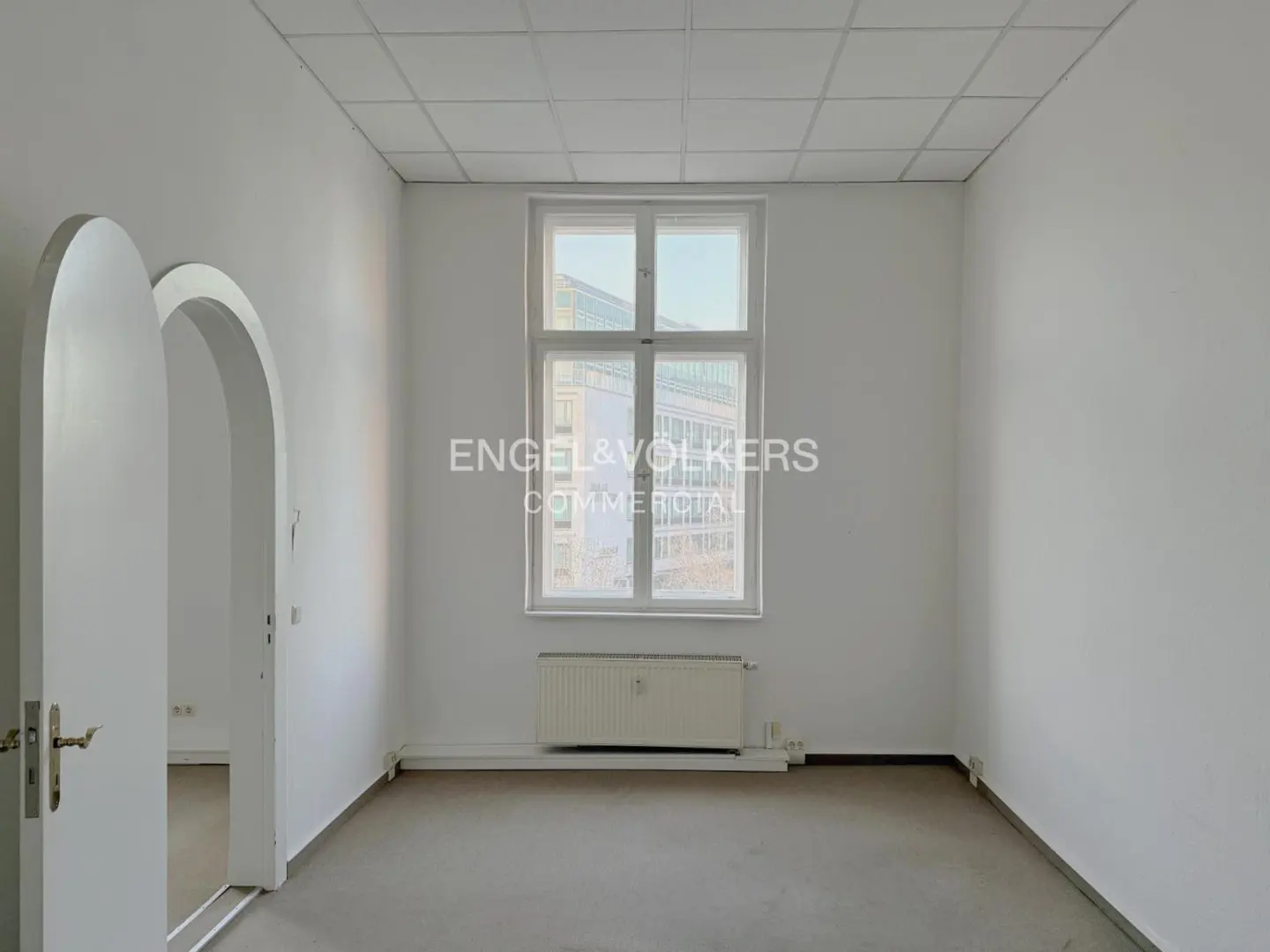 Empty white room with a window, radiator, and arched doorway. Engel & Volkers logo is superimposed on the window.