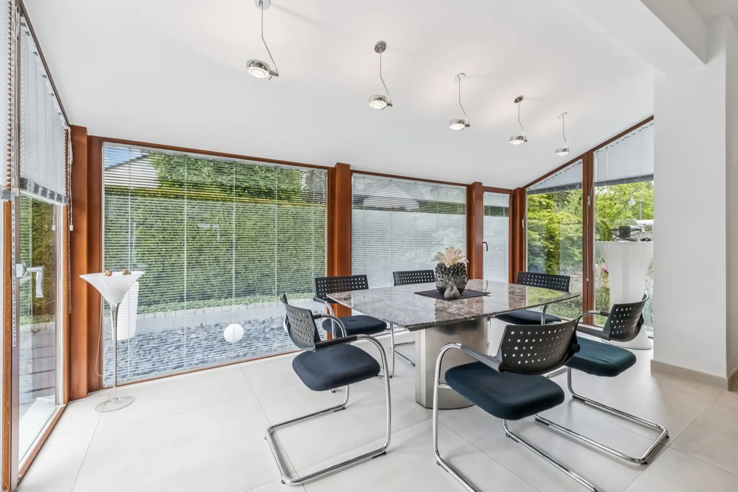 Bright sunroom with a marble table, black chairs, and large windows overlooking a green garden. Modern pendant lights hang from the white ceiling.
