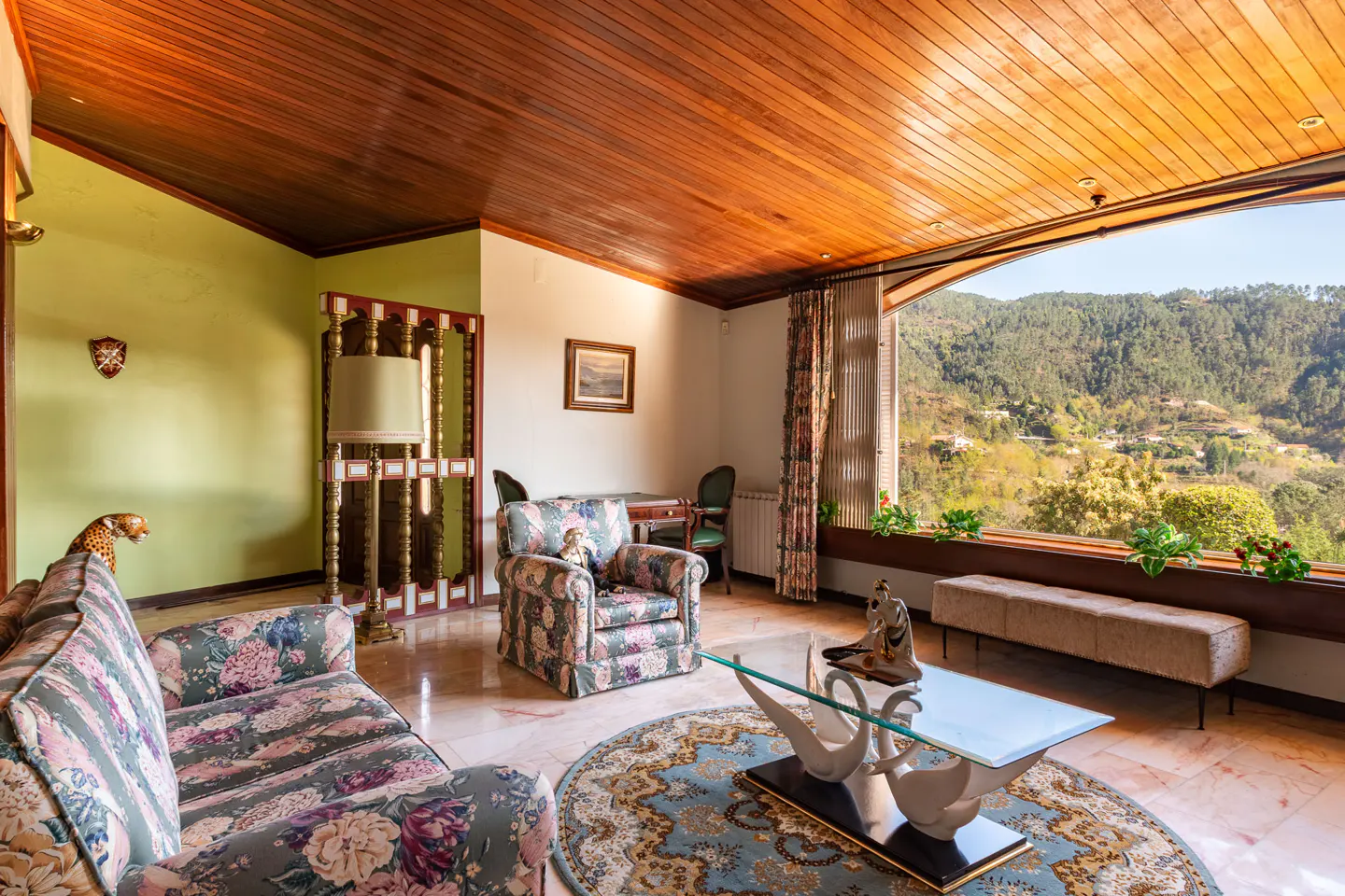 Living room with floral sofa and chair, round rug, and swan coffee table. Large window with mountain view. Wood ceiling and marble floor.