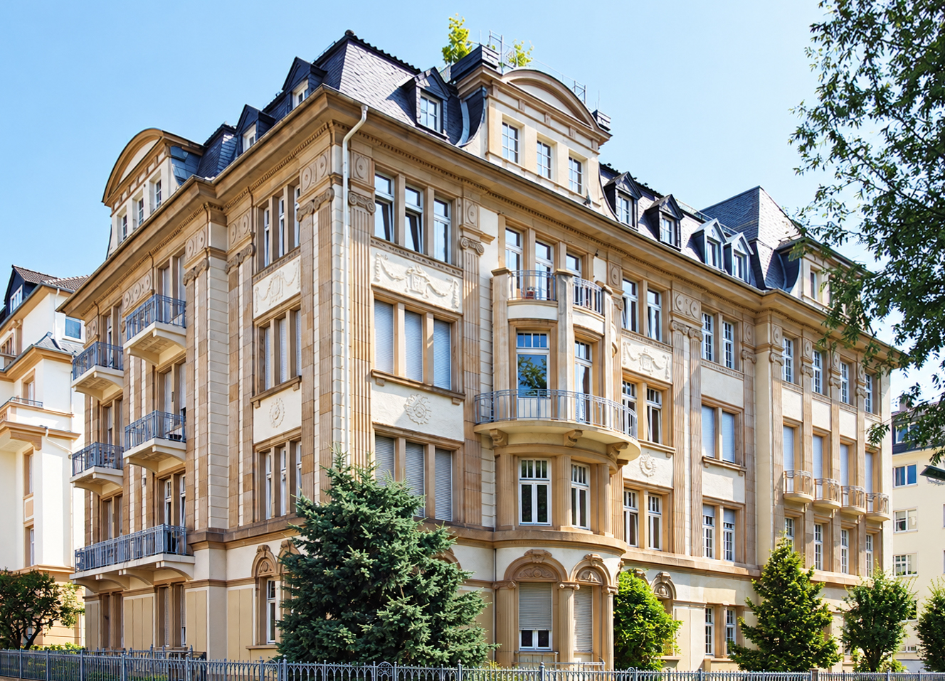 A grand, multi-story beige stone apartment building with ornate architectural details, balconies, and a dark slate roof. [AI-generated text]