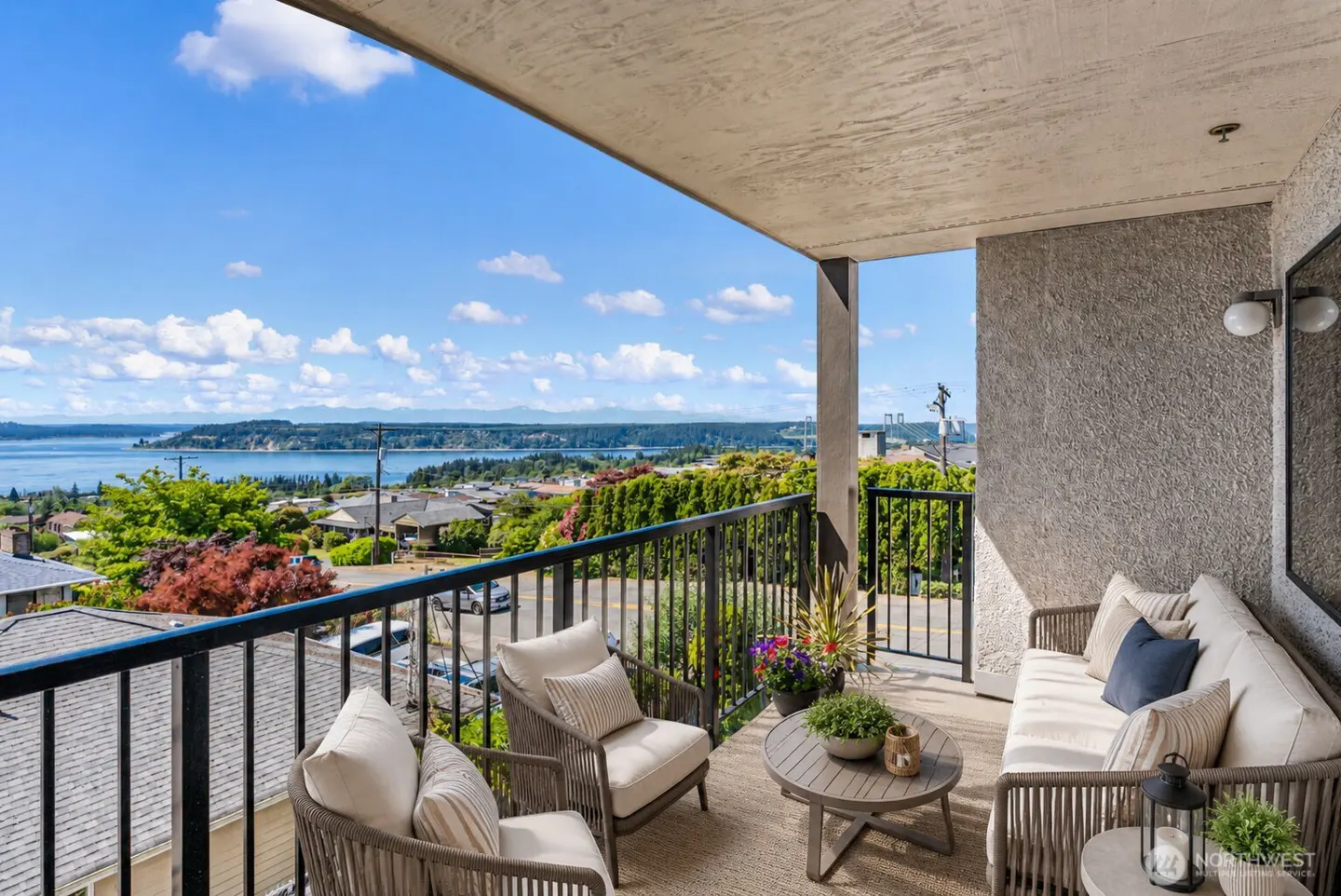 Covered balcony with outdoor seating, a round table, and a scenic view of the water, trees, and blue sky.