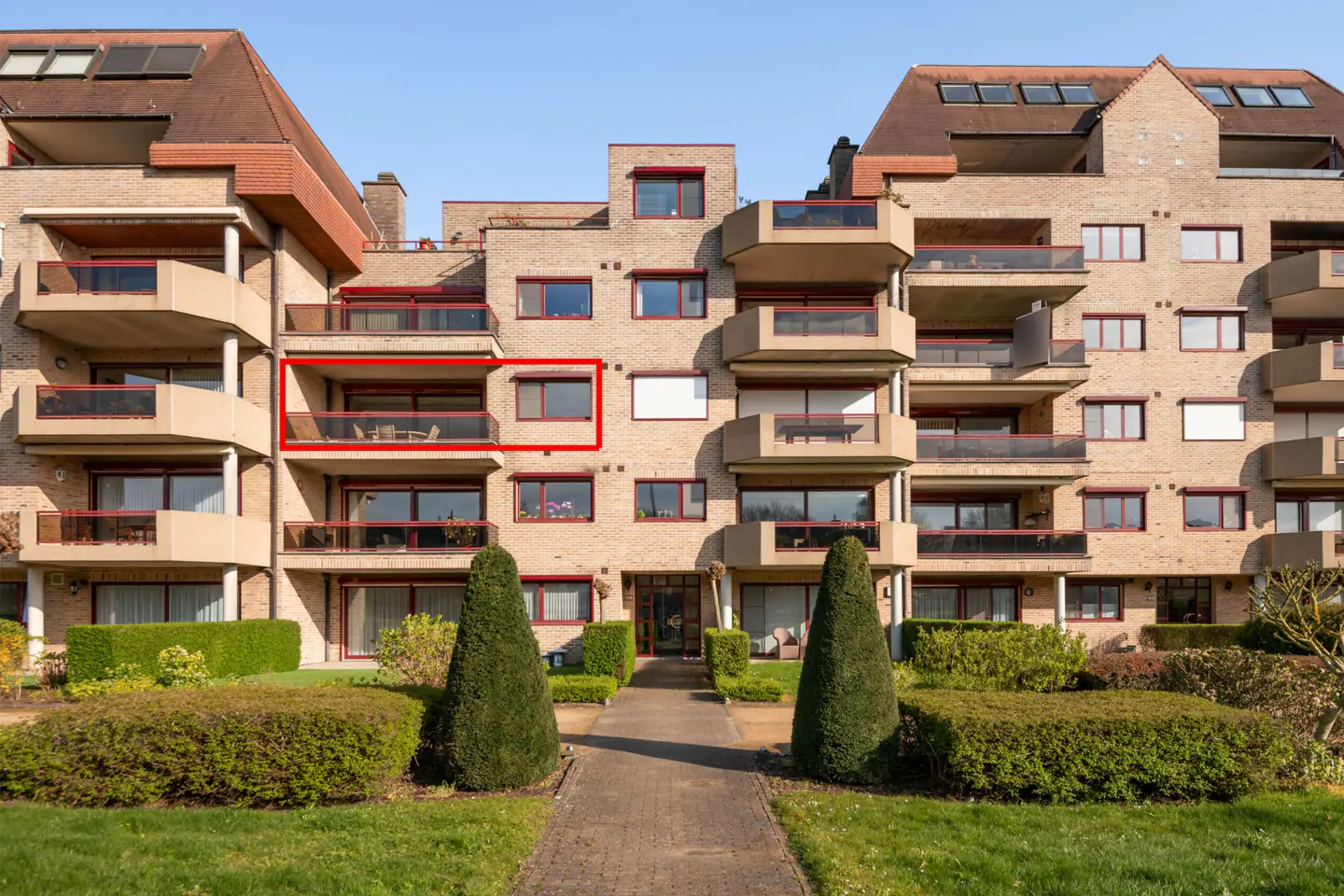 Exterior of a tan brick apartment building with balconies and a red-framed unit. A stone path leads through manicured green bushes.