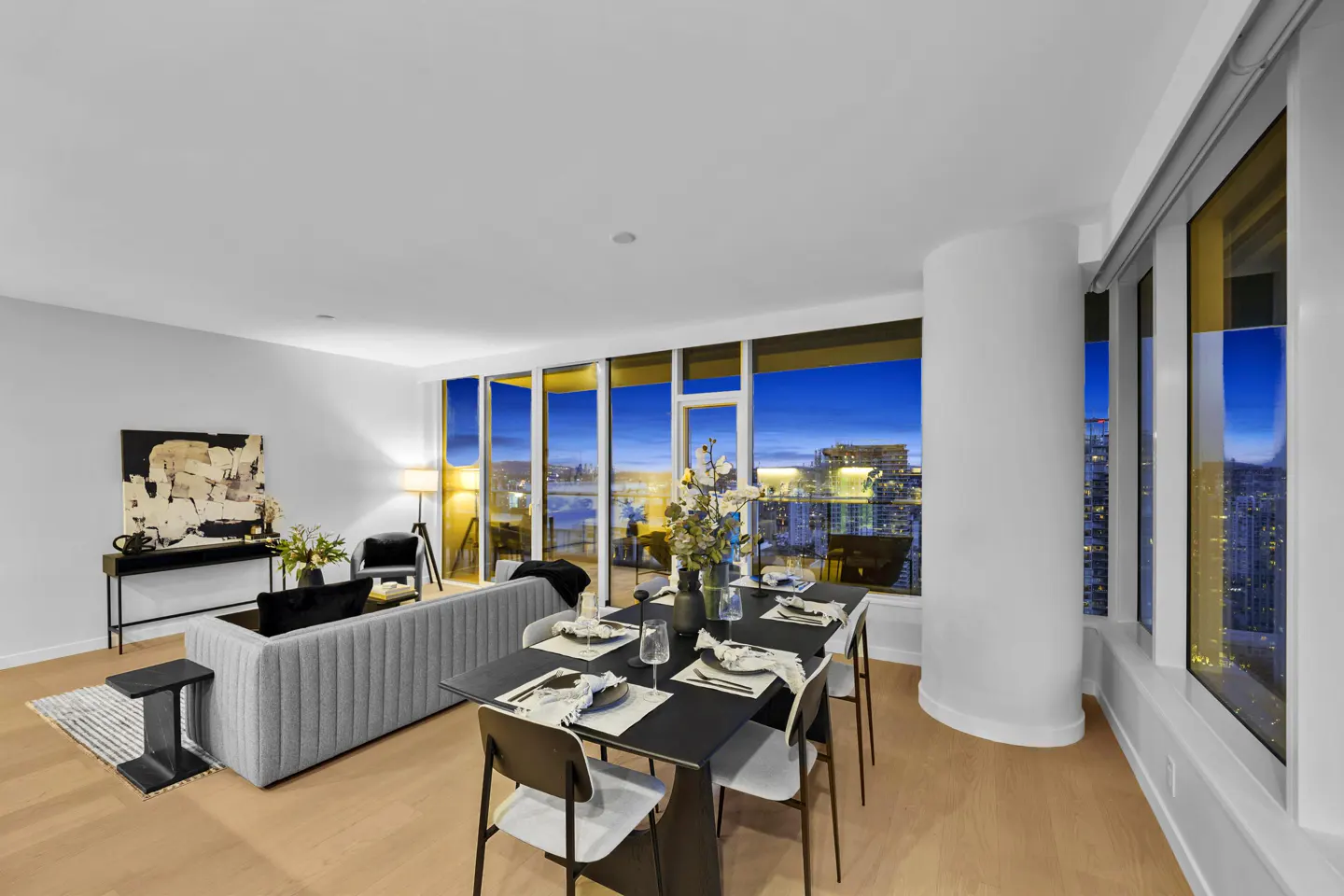Bright, modern condo interior with a gray sofa, black dining table set for six, and a stunning city view at dusk.