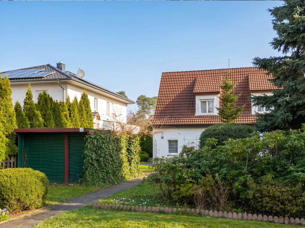 A suburban scene with two houses, one white with solar panels, the other with a red tile roof, surrounded by green trees and bushes.