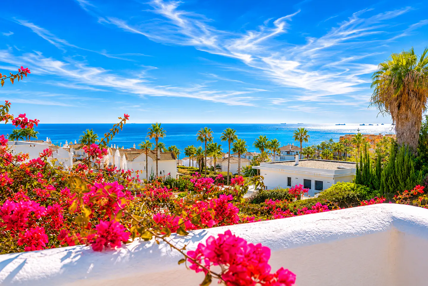 View from a white balcony with pink bougainvillea overlooking white buildings, palm trees, and the blue ocean under a bright blue sky.