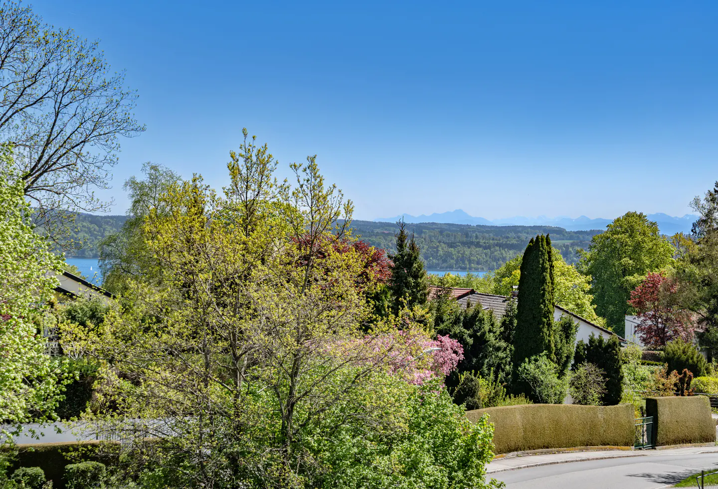 Scenic view of a lake and mountains beyond lush green trees and rooftops under a clear blue sky.