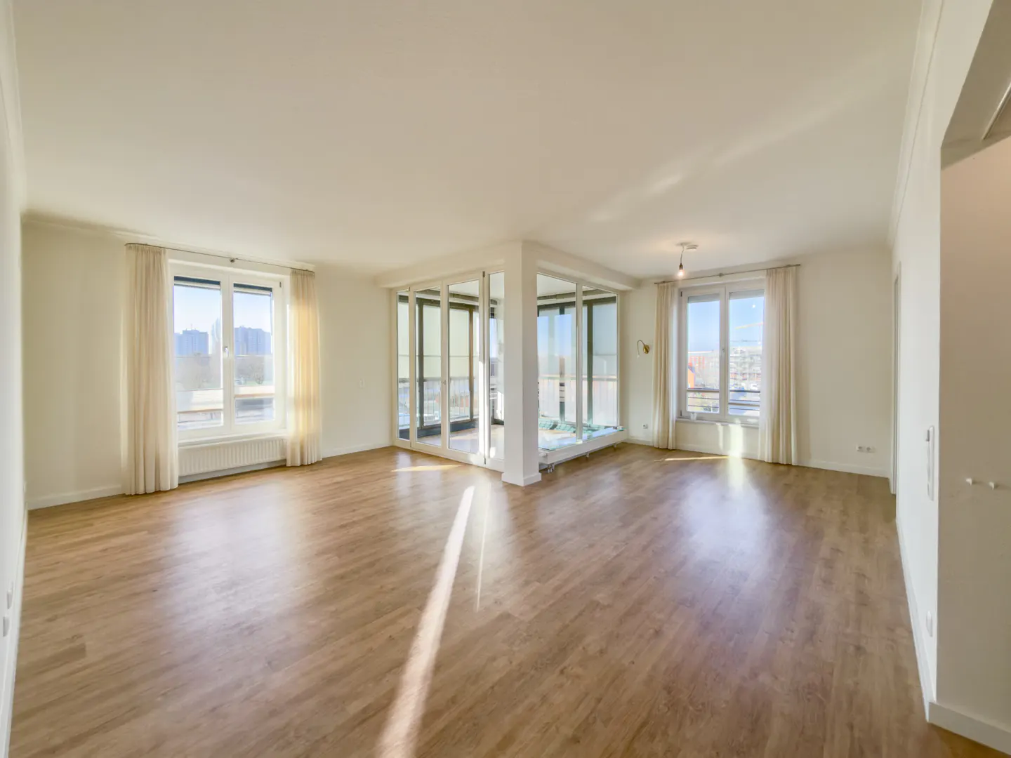 Bright, empty room with wood floors, white walls, and large windows with sheer curtains. A glass-enclosed balcony area is in the center.