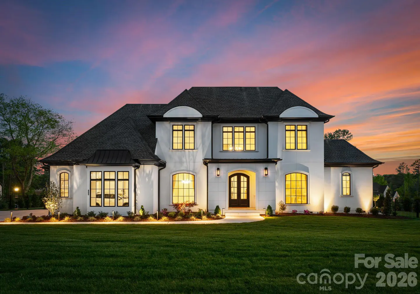 A white two-story luxury home with dark trim and glowing interior lights at sunset, set against a vibrant orange sky.