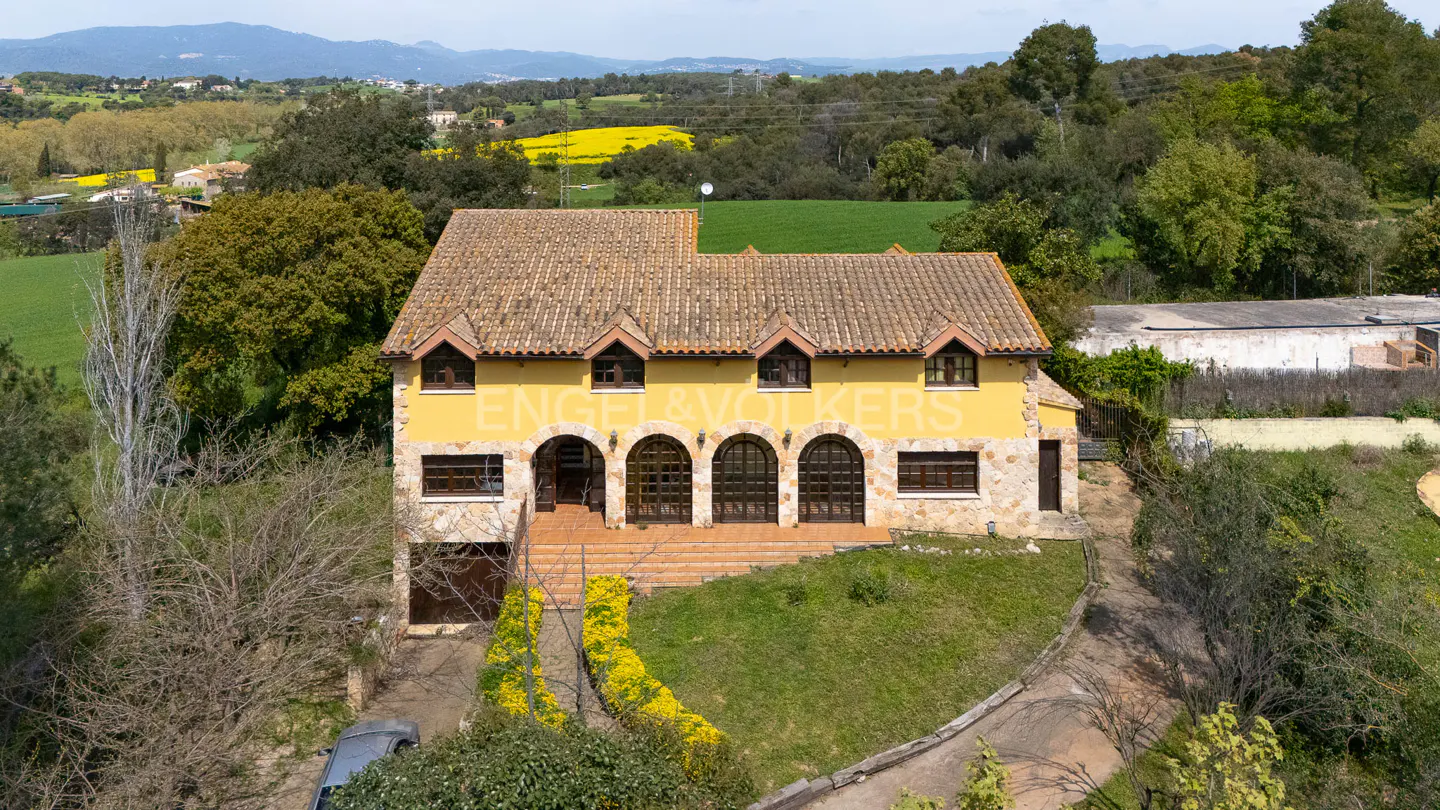 Two-story yellow house with a brown tile roof and arched windows. The house is surrounded by green grass and trees. In the background, there are green fields and mountains.