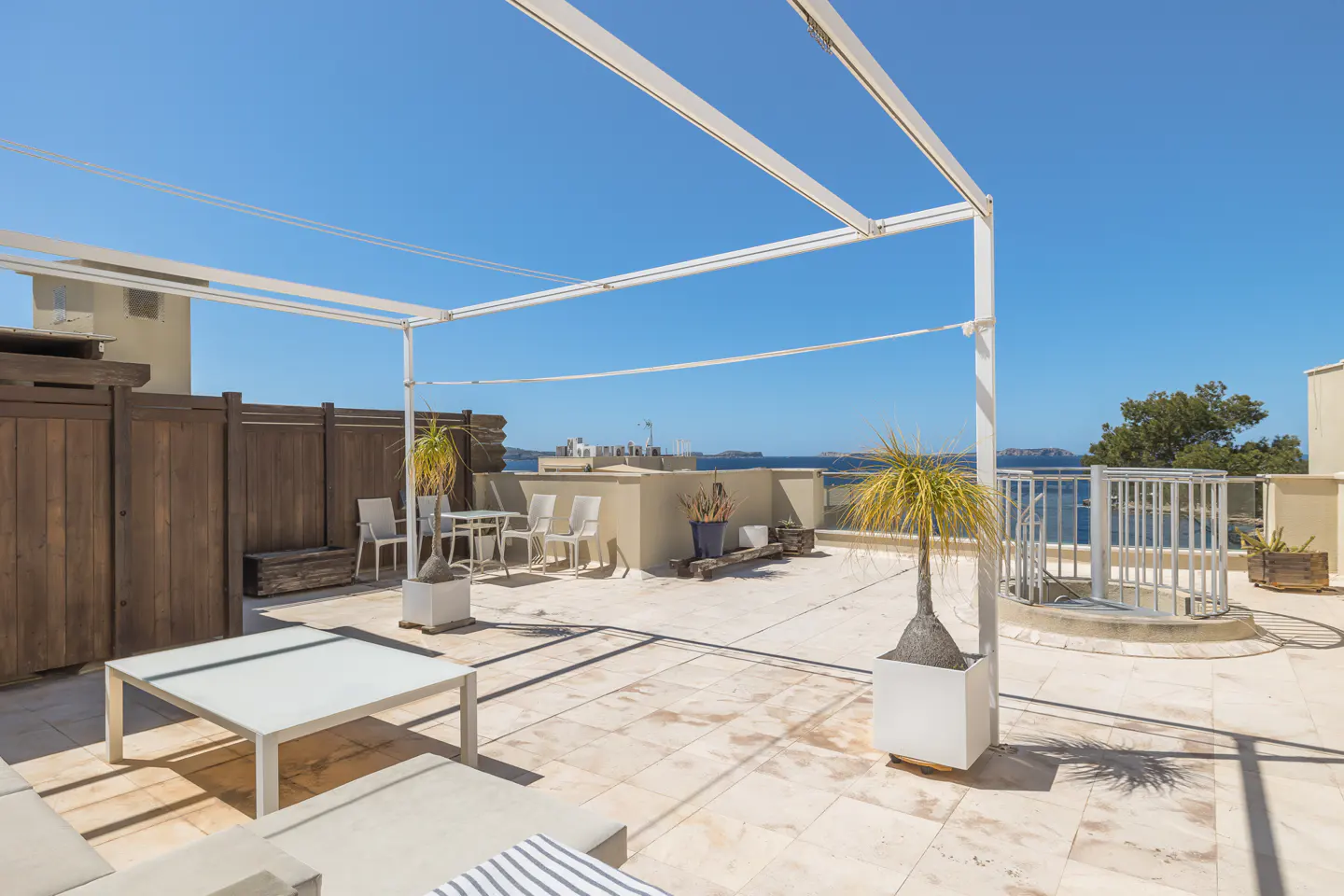 Rooftop patio with white pergola, table, chairs, and potted plants. Ocean view in the background under a clear blue sky.
