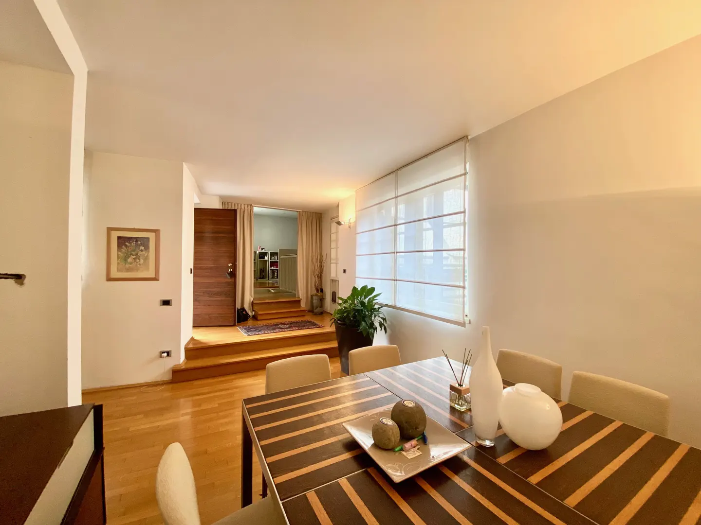 A dining room with a striped table, beige chairs, and white walls. A doorway with wooden steps leads to another room. A large window with blinds is on the right.