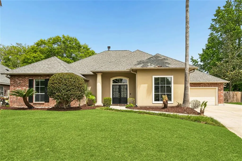 A single-story home with a manicured lawn, tan stucco, brick accents, and a gray roof under a clear blue sky.