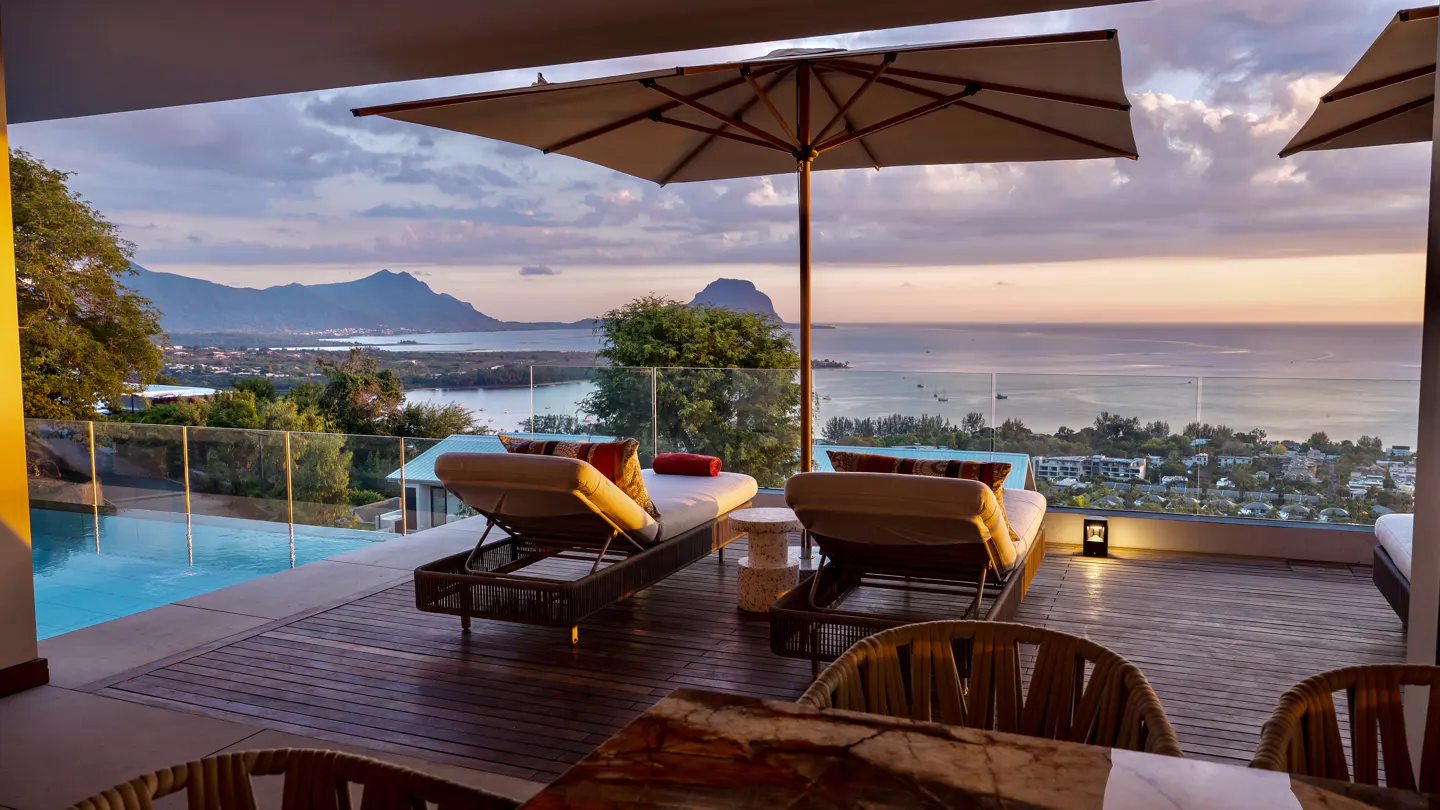 Luxury patio with two lounge chairs, umbrella, and ocean view at sunset. Mountains in the background.