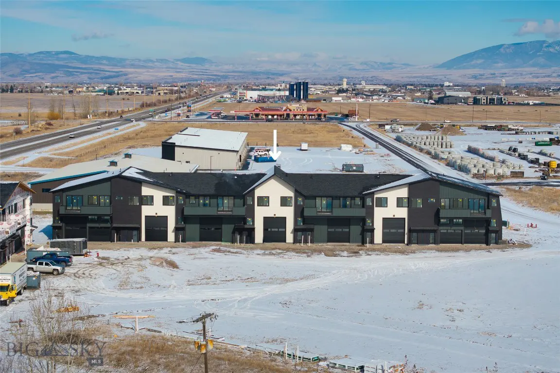 Modern townhouses with black garage doors and gray siding stand in a snowy field, with mountains and a city skyline in the background.