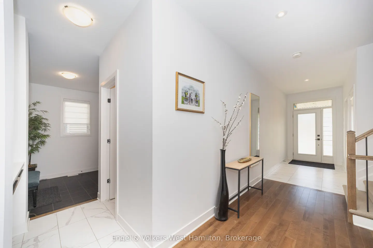 Bright foyer with white walls, wood floors, and a console table with a tall black vase. A framed picture hangs on the wall. Front door visible.