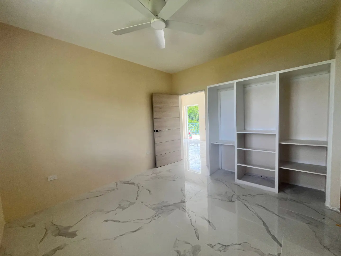Bedroom with marble floors, beige walls, and a white ceiling fan. An open door leads to a bright outdoor area. Built-in white shelving unit.