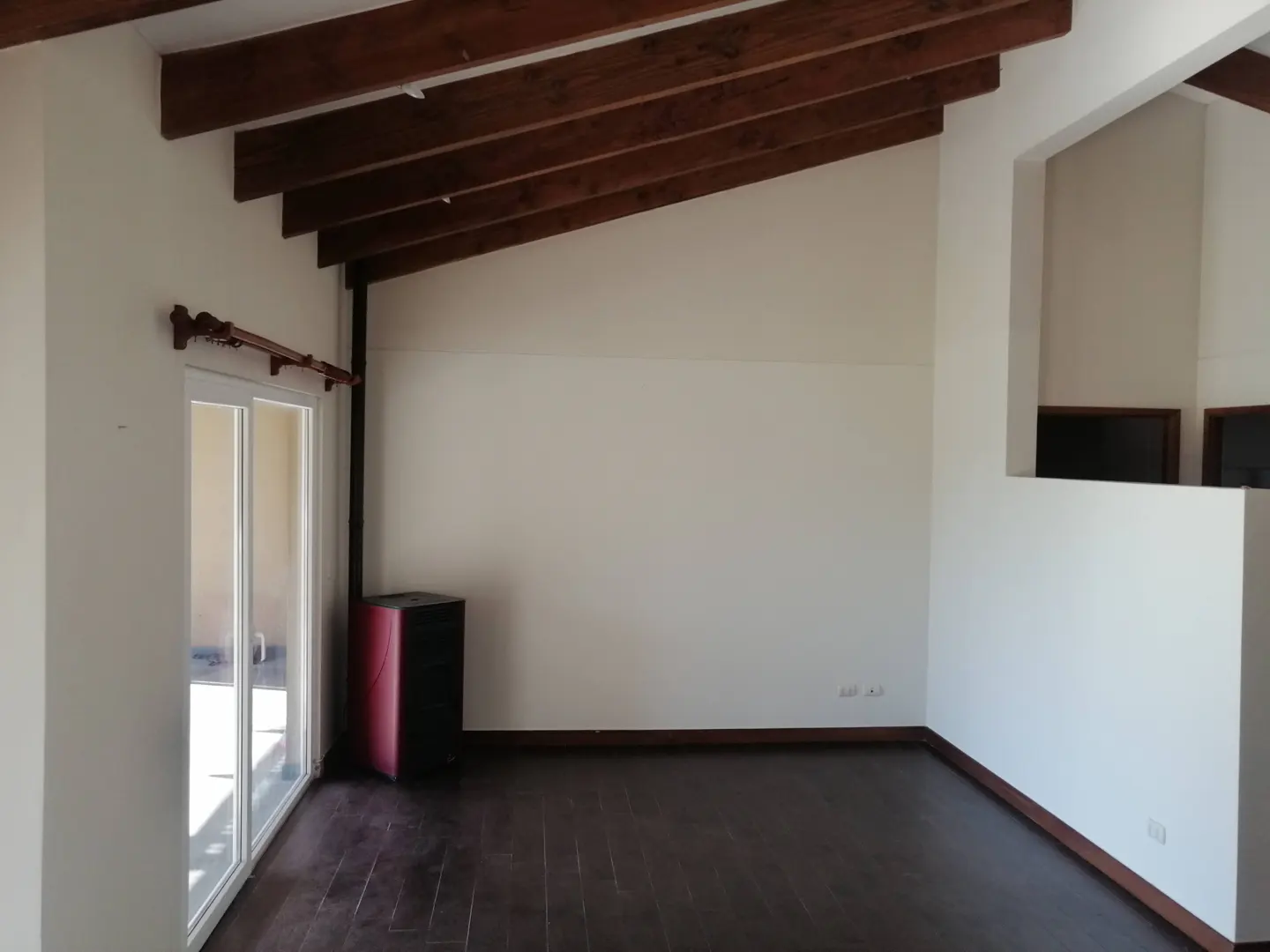 Empty room with dark wood floors, white walls, and exposed beams. A sliding glass door leads to a patio. A red and black heater sits near the wall.