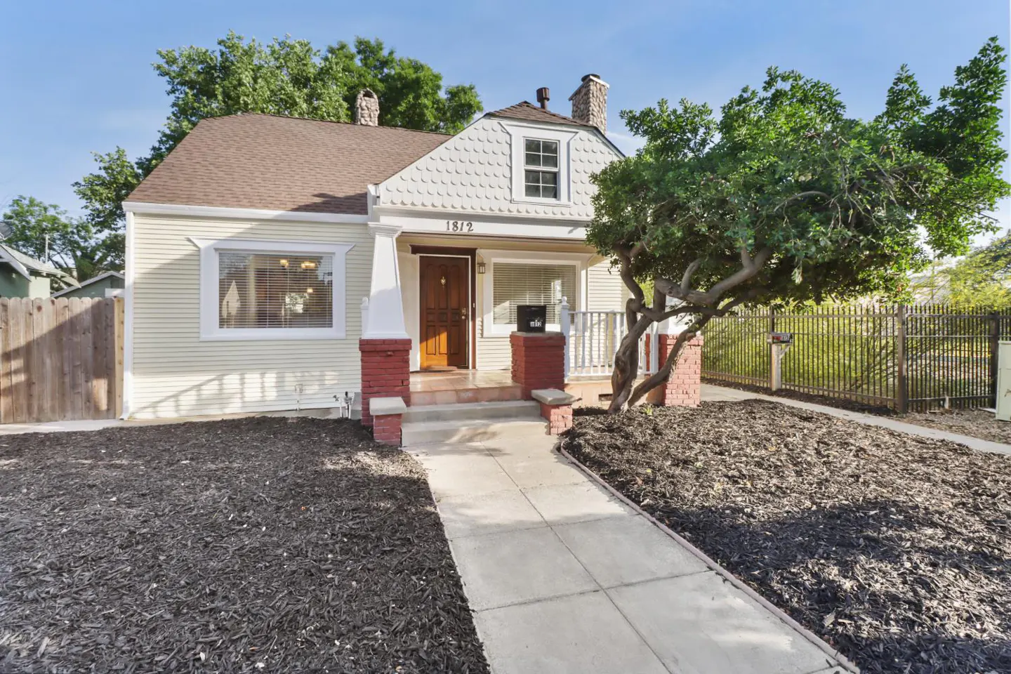 A quaint, light yellow house with a brown roof and a concrete walkway leading to the front door. A large tree stands to the right.