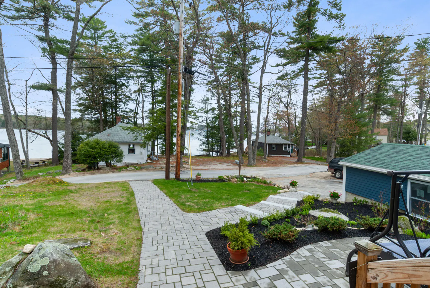 View of a lake house with a gray brick walkway, green lawn, and tall trees. A blue house with a green roof is on the right.