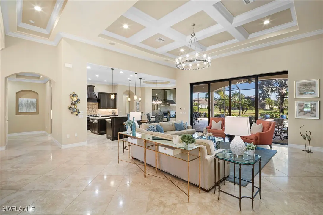 Open-concept living room with beige walls, tile floors, and coffered ceiling with a chandelier. Kitchen in background.