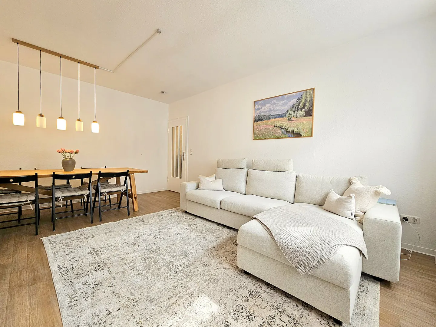 Bright living room with a beige sectional sofa, a wooden dining table with black chairs, and a patterned rug on a wood floor. A landscape painting hangs on the white wall.