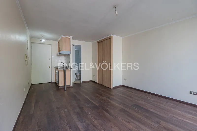 A bright studio apartment with dark wood floors, a small kitchen, and a bathroom. The walls are painted in a light neutral color.