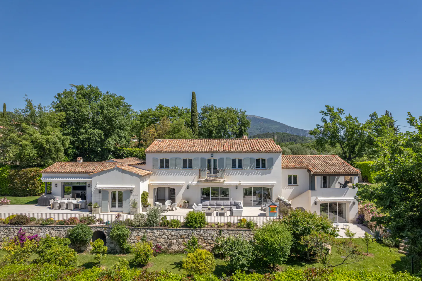 Exterior view of a two-story white house with a terracotta roof and blue shutters, surrounded by lush greenery.