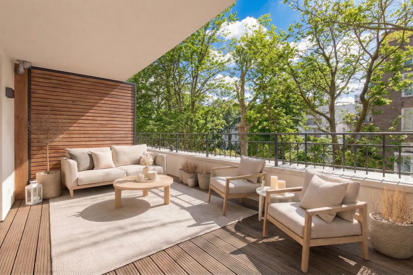 A sunlit balcony with a beige sofa, two chairs, and a round table on a light rug. Green trees are visible beyond the black railing.