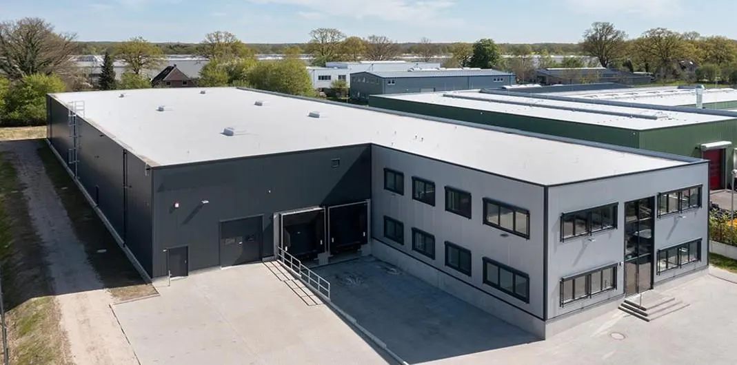 Aerial view of a modern, gray industrial building with a white roof and loading docks. Trees are visible in the background.